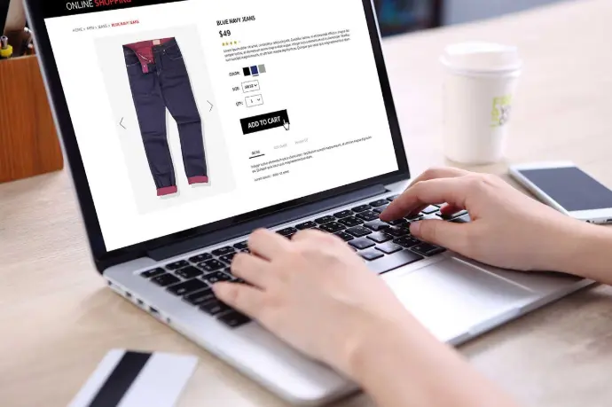 A person using a laptop to shop online, viewing a product page for jeans and adding them to the cart